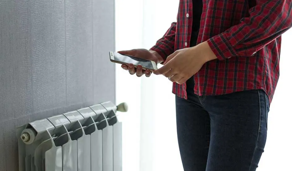 Un mur gris jouxte un radiateur blanc. Une personne en chemise à carreaux rouge tient un smartphone.