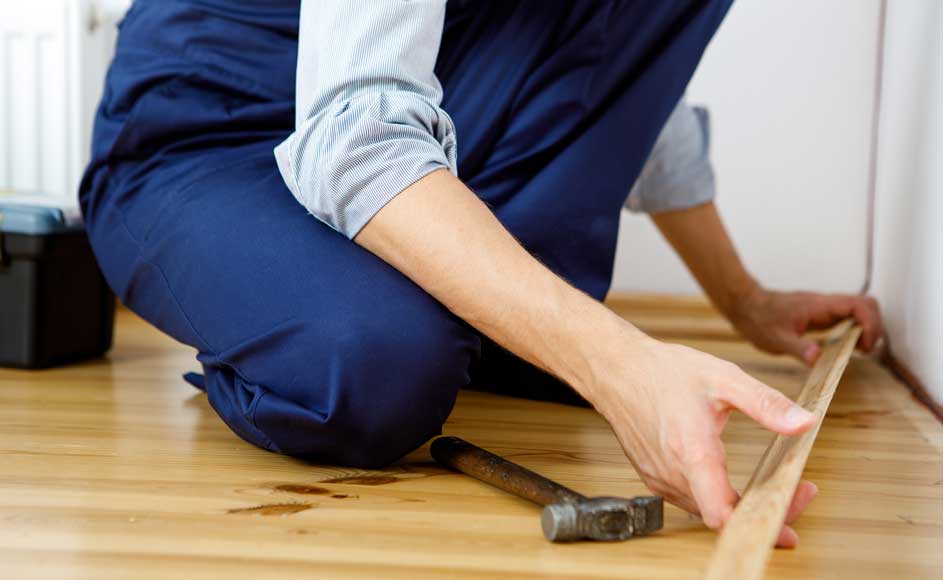 Een persoon in blauwe kledij knielt op een houten vloer naast een witte muur. Een hamer en een gereedschapskist zijn zichtbaar.