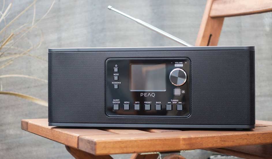 Een zwarte radio met antenne staat op een houten stoel tegen een grijze achtergrond.