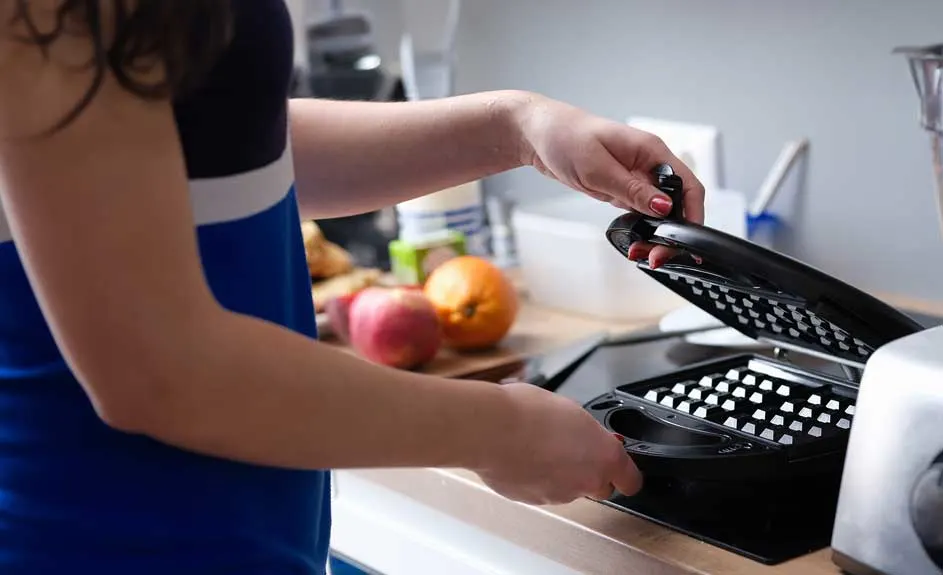 Une gaufrier noir est ouvert sur un comptoir de cuisine avec des fruits en arrière-plan.