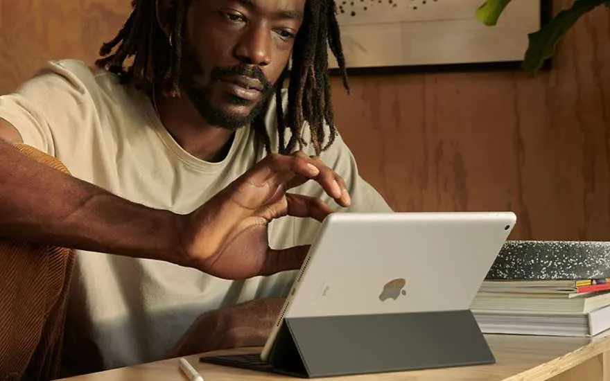 Een zilverkleurige iPad staat op een houten tafel met een donkergrijs toetsenbordhoes. Een witte stylus ligt naast het apparaat.