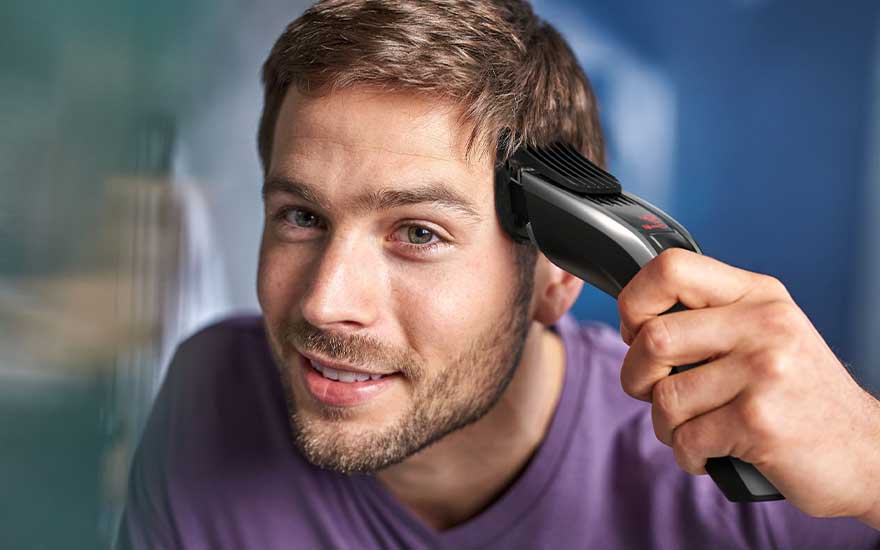 Een glimlachende man trimt zelf zijn haar met een elektrische tondeuse, rustig in de spiegel kijkend in een ontspannen badkamer.