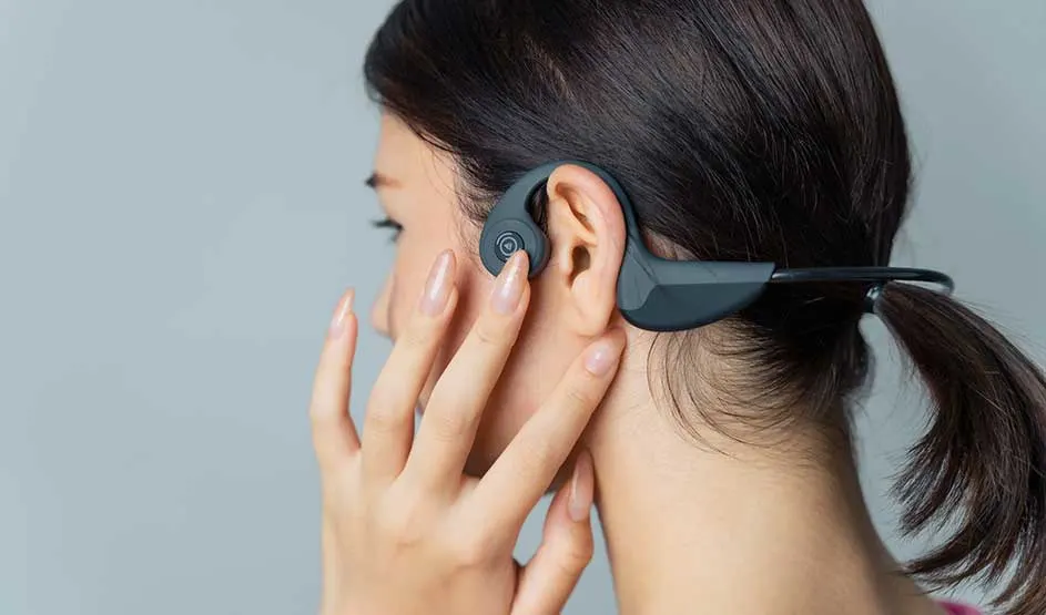 Vrouw met staart draagt zwarte bone‑conduction koptelefoon achter haar oor en raakt zacht de knop aan, voor een effen grijze achtergrond.