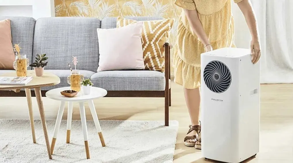Een witte airconditioner staat in een woonkamer met een grijze zetel, houten tafels en een wit tapijt.
