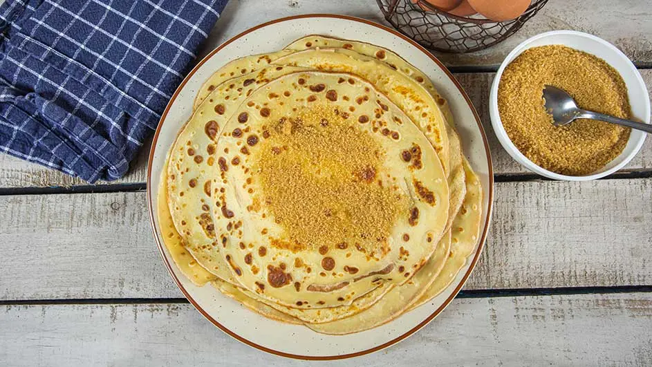 Pannenkoeken op een bord met bruine suiker. Een blauw-wit geruite doek en een kom suiker staan op een houten tafel.