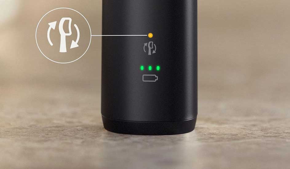 Zwarte cilinder met wit rotatiesymbool, gele oplaadindicator en groene batterijstatus-LED's. Staat op een beige oppervlak.