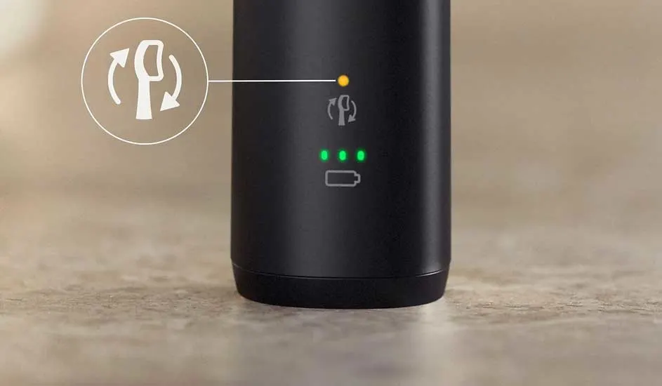 Zwarte cilinder met wit rotatiesymbool, gele oplaadindicator en groene batterijstatus-LED's. Staat op een beige oppervlak.