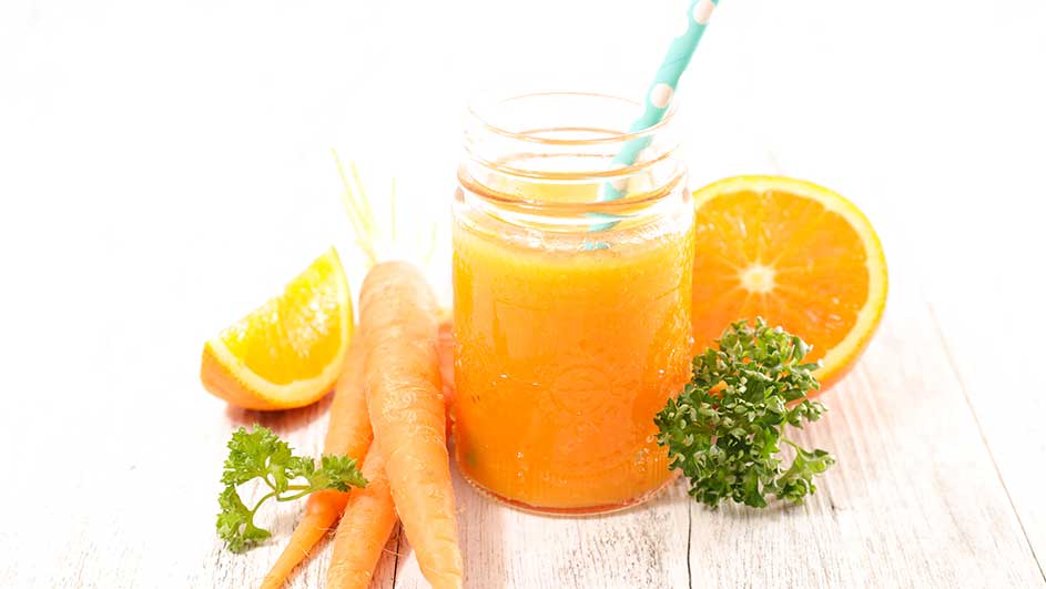 Un verre de jus orange avec une paille bleue, des carottes, des oranges et du persil sur une table en bois blanche.