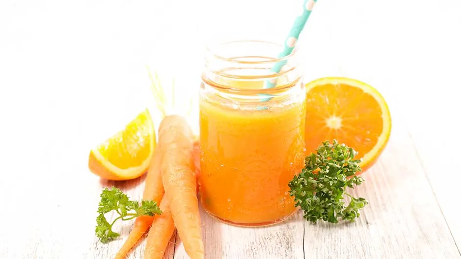 Un verre de jus orange avec une paille bleue, des carottes, des oranges et du persil sur une table en bois blanche.