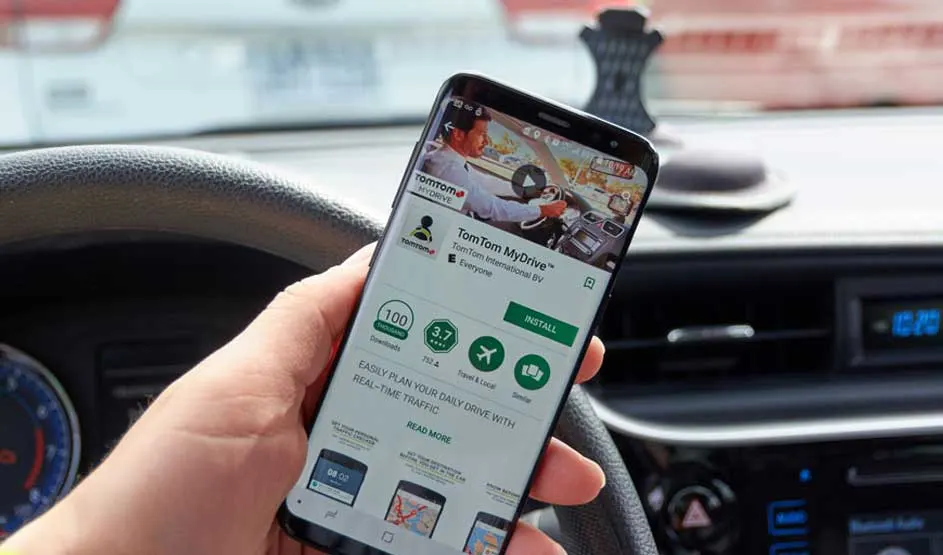 Een hand houdt een smartphone met de TomTom MyDrive app in een auto, stuur en dashboard zijn zichtbaar.