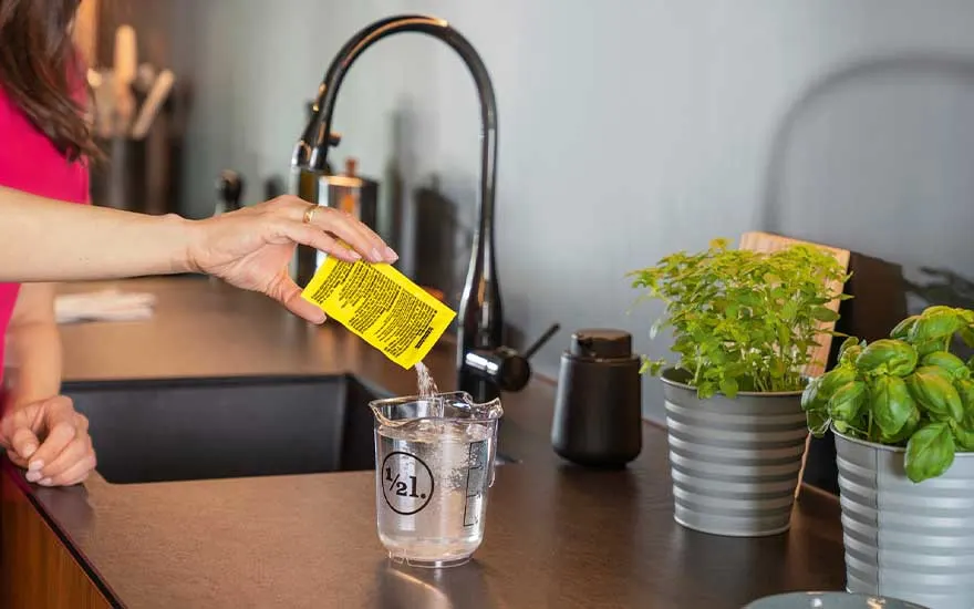 Hand giet geel zakje in maatbeker met water. Aanrecht met kraan, kruiden in potten en bruine zeepdispenser.
