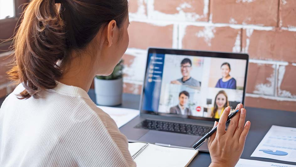 Femme devant un ordinateur portable en appel vidéo, prenant des notes. Mur de briques en arrière-plan.