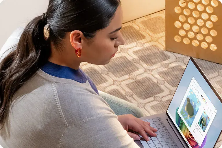 Een vrouw zit op de grond met een laptop en bewerkt een kleurrijke afbeelding, naast een kartonnen doos met ronde uitsparingen.