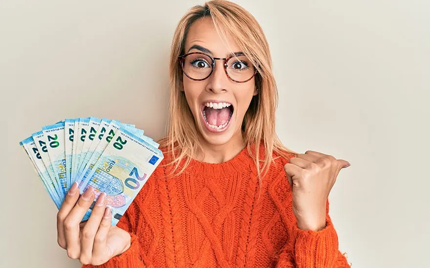 Une femme tient des billets de 20 euros, sourit et lève le poing devant un fond beige.