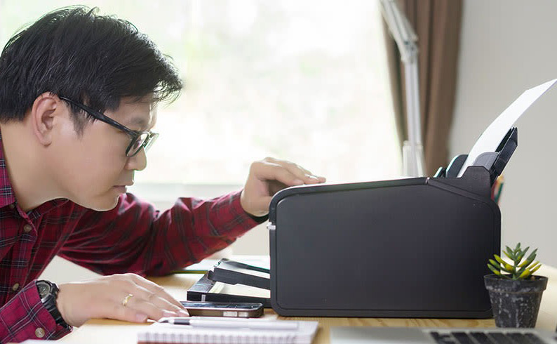 Un homme utilise une imprimante noire sur un bureau avec un ordinateur portable, un bloc-notes et une plante en pot.