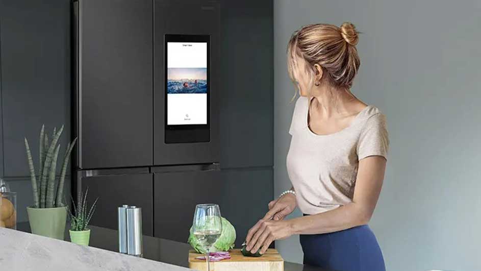 Frigo gris foncé avec écran affichant une scène de plage. Un comptoir présente des plantes, des couverts, un verre et un chou coupé.