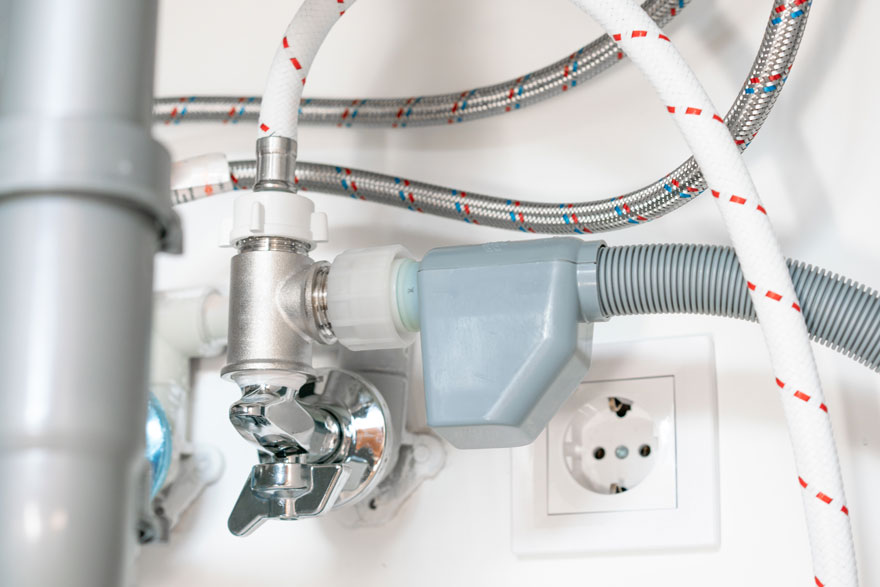 Een close-up van een gootsteenafvoer met zilverkleurige buizen, witte kabels met rode accenten, een grijze aansluitdoos en een wit stopcontact.