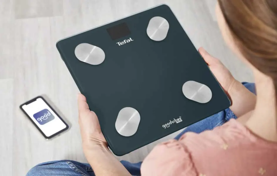 Een persoon houdt een zwarte Tefal-weegschaal vast met vier metalen sensoren. Naast de weegschaal ligt een smartphone met een gezondheidsapp op het scherm.