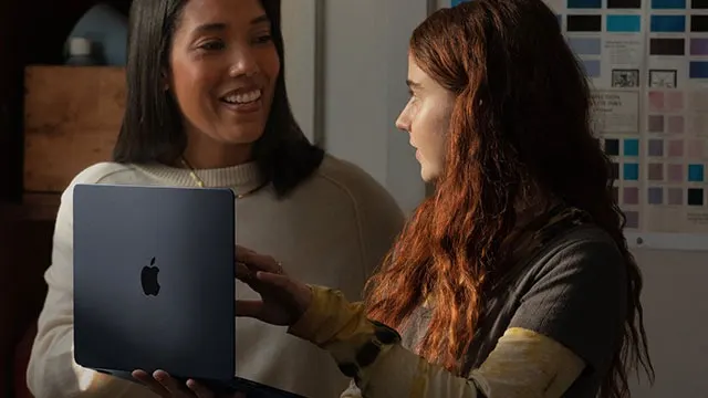 Twee mensen kijken naar een zwarte laptop met een Apple-logo. Achtergrond toont een muur met kleurstaaltjes.