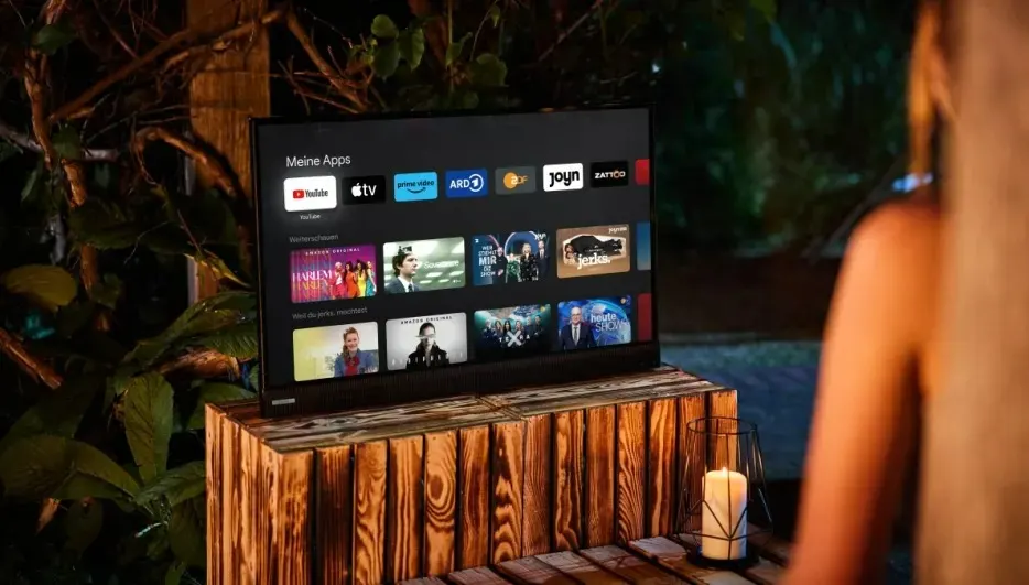 Zicht op een tv op een houten kast met apps op het scherm, buiten bij groen en een kaars.