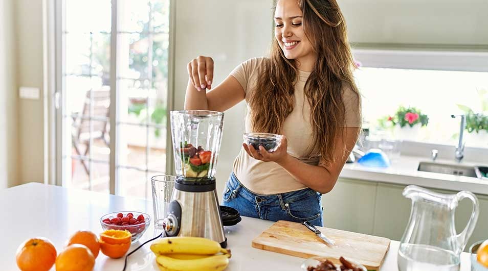Op een wit aanrecht staat een blender met fruit. Sinaasappels, bananen, en bessen liggen ernaast. Een glazen kan staat achteraan.
