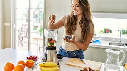 Op een wit aanrecht staat een blender met fruit. Sinaasappels, bananen, en bessen liggen ernaast. Een glazen kan staat achteraan.