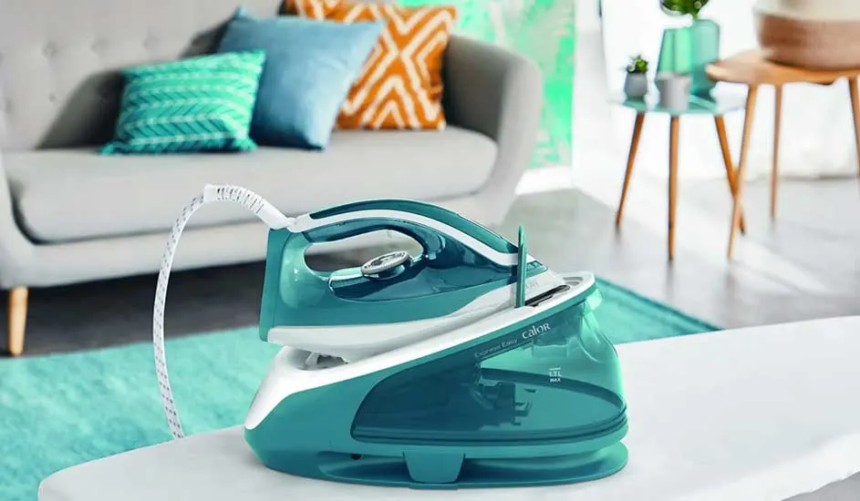 Een turkoois strijkijzer met basisstation staat op een witte ondergrond, met een bank en tafels op de achtergrond.