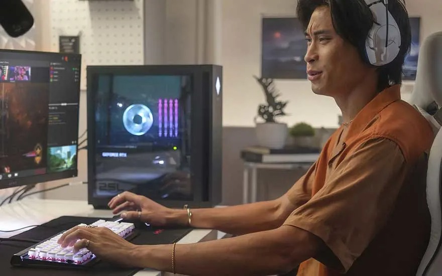 A gamer wearing a headset sits at a desk, typing on a lit keyboard and watching monitors; a PC with RGB lighting sits beside him.