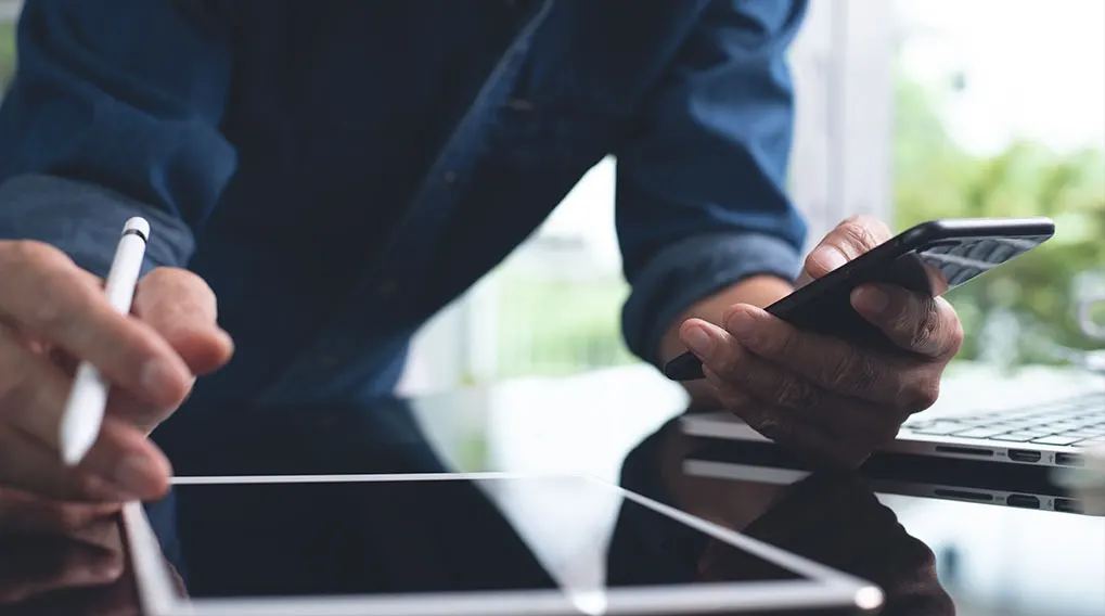 Een persoon gebruikt een witte stylus op een tablet en houdt een zwarte smartphone vast, zittend aan een tafel met een laptop.