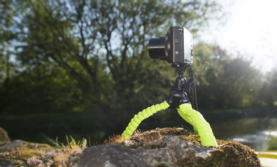 Een zwarte camera op een groen statief staat op een rots, met bomen en water op de achtergrond.