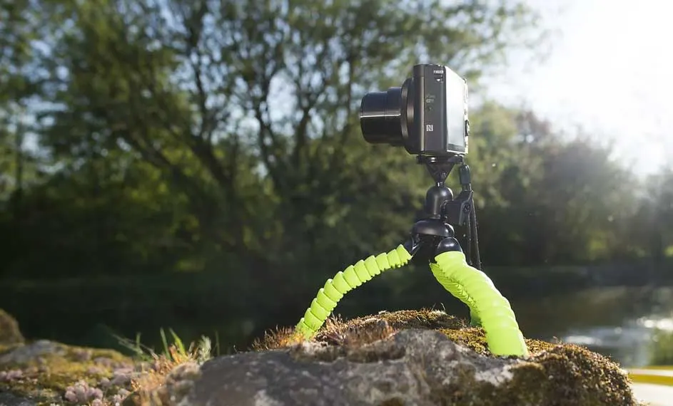 Een zwarte camera op een groen statief staat op een rots, met bomen en water op de achtergrond.