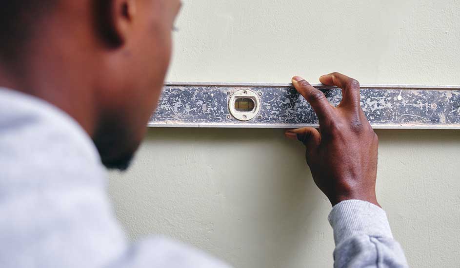 Un niveau à bulle argenté est tenu contre un mur beige. Une main ajuste l'outil.