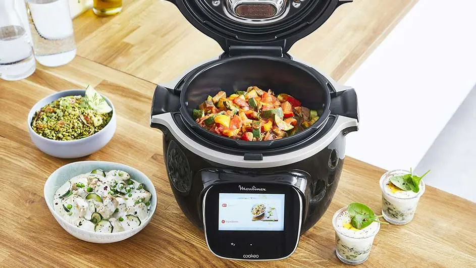 Een zwarte multicooker staat op een houten tafel, met erin een gerecht. Er staan ook kommen met eten en glazen.