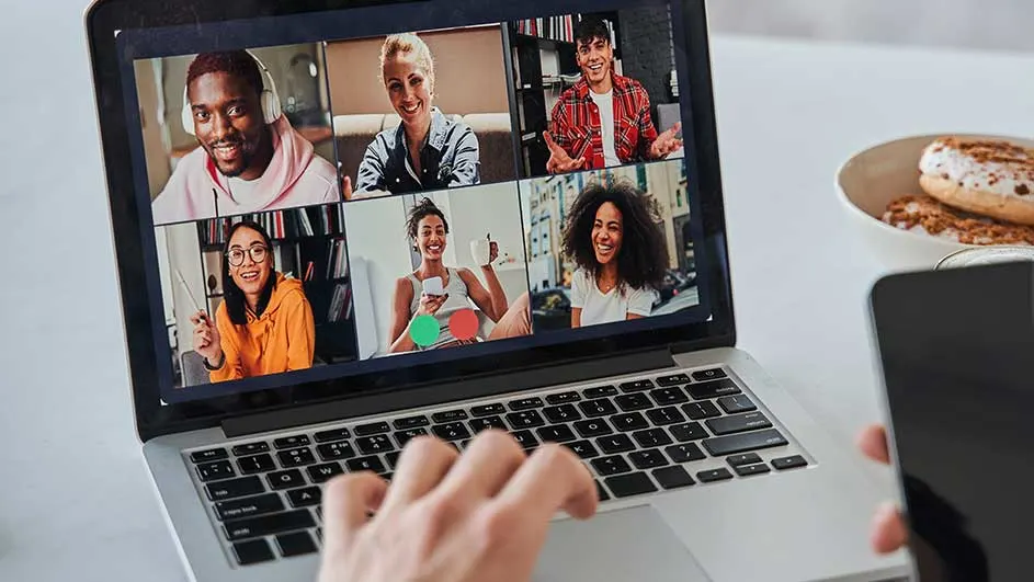 Laptop toont een videogesprek met zes deelnemers. Een hand typt op het toetsenbord. Een telefoon en gebak staan op tafel.