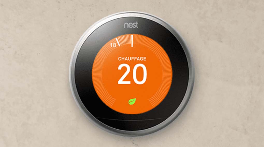 Thermostat Nest rond sur un mur beige. L'écran affiche "CHAUFFAGE 20" en blanc sur fond orange, avec une petite feuille verte.