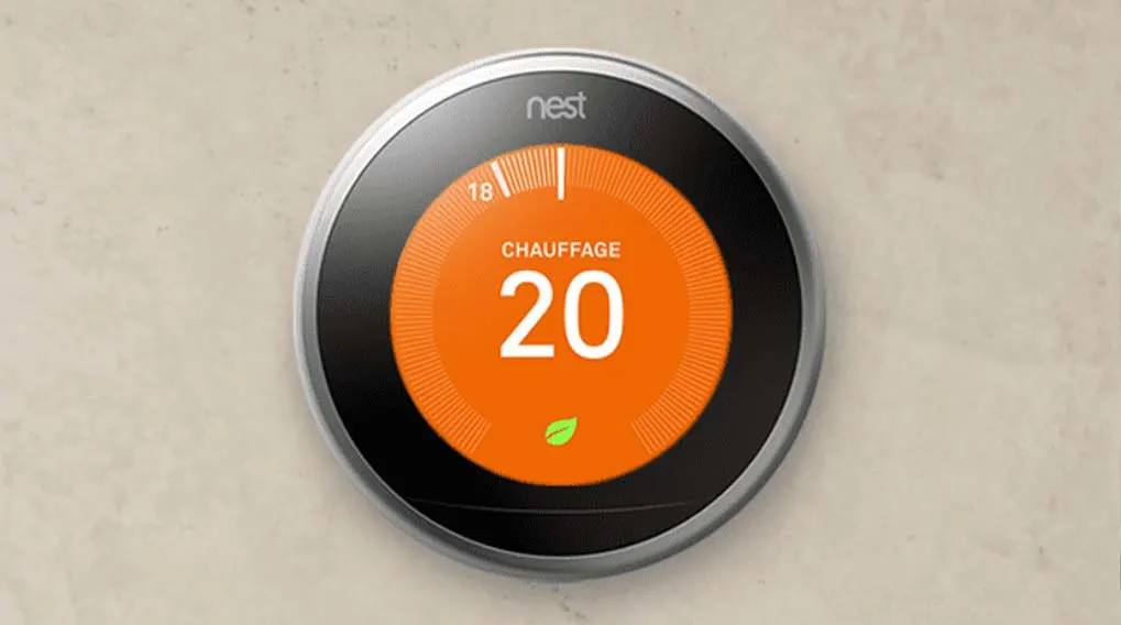 Thermostat Nest rond sur un mur beige. L'écran affiche "CHAUFFAGE 20" en blanc sur fond orange, avec une petite feuille verte.