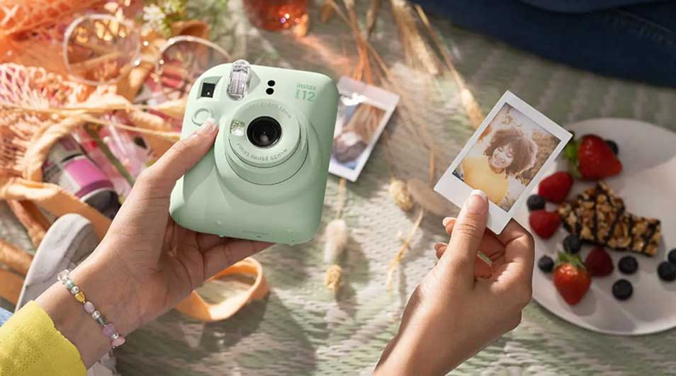 A person is holding a mint green Fujifilm Instax camera and looking at a freshly made Polaroid photo, with fruit and snacks in the background.