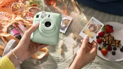A person is holding a mint green Fujifilm Instax camera and looking at a freshly made Polaroid photo, with fruit and snacks in the background.