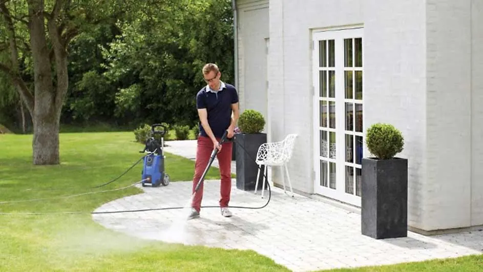 Een hogedrukreiniger spuit water op een terras naast een wit gebouw met een deur en groene planten.