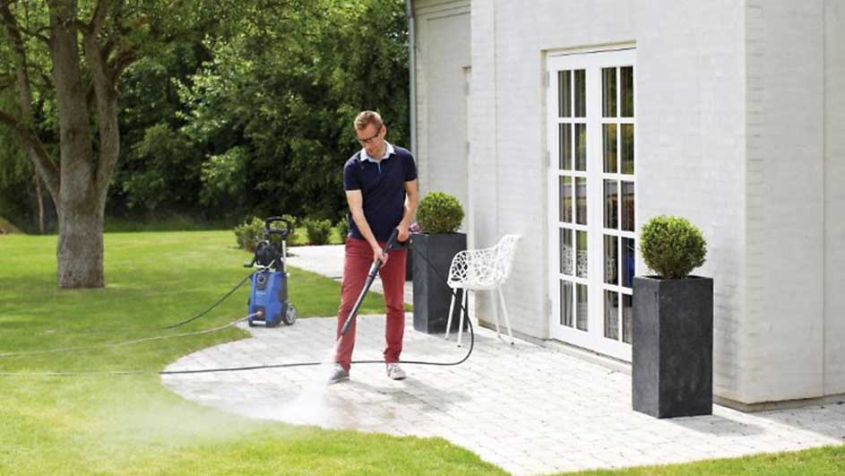 Un homme utilise un nettoyeur haute pression sur une terrasse près d'une maison blanche avec jardin et arbres.