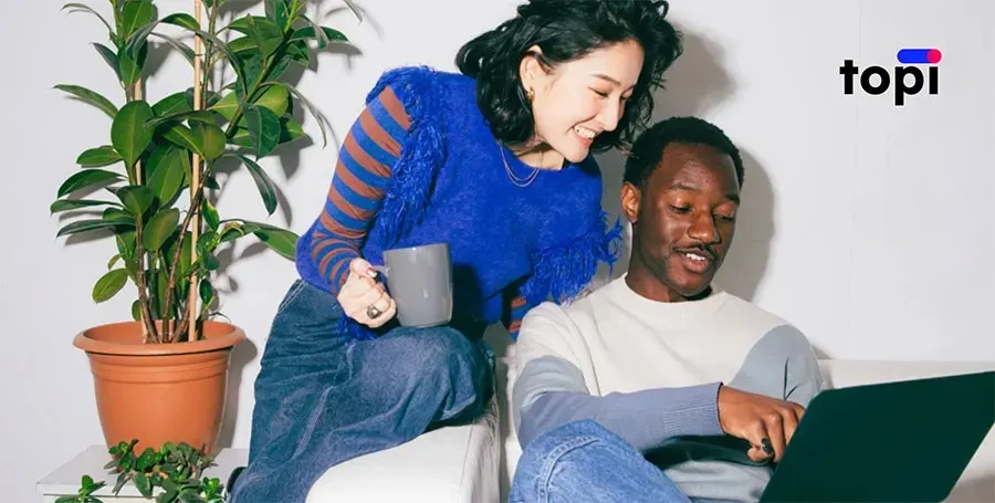 Deux collègues se détendent dans une pièce lumineuse; elle se penche avec une tasse, souriante, tandis qu’il lui montre quelque chose sur son ordinateur portable.