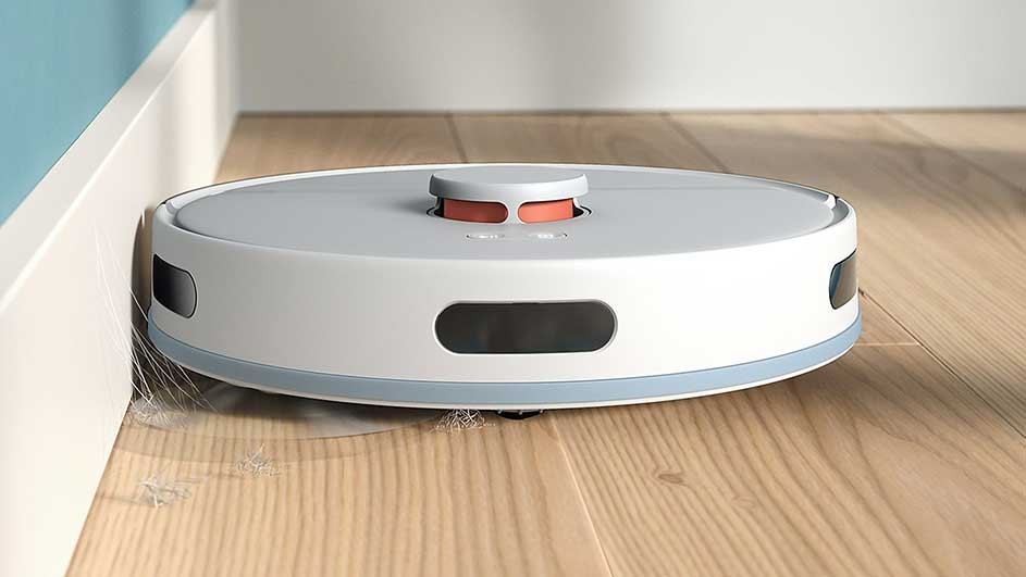Un aspirateur robot blanc et gris nettoie un plancher de bois près d'une plinthe blanche.
