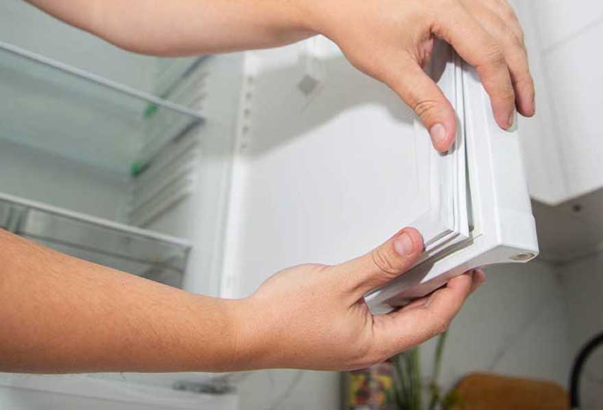 Deux mains installent soigneusement un joint blanc autour de la porte d’un réfrigérateur ouvert. L’intérieur vide, les clayettes en verre et la porte sont visibles.