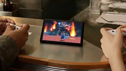 Een Nintendo Switch-console staat op een metalen oppervlak, met een videospel erop. Twee personen houden controllers vast.
