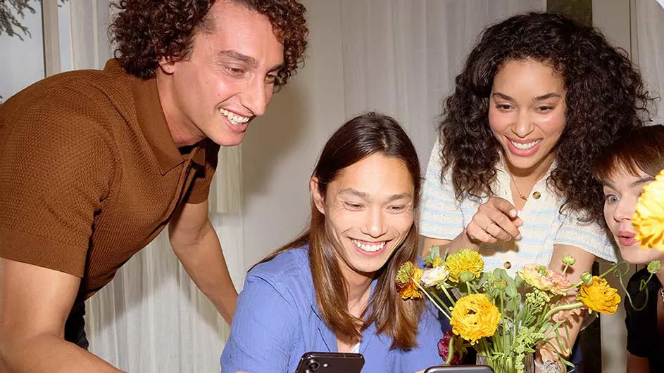 Quatre personnes regardent un téléphone portable près d'un bouquet de fleurs jaunes et orange. Rideaux blancs en arrière-plan.