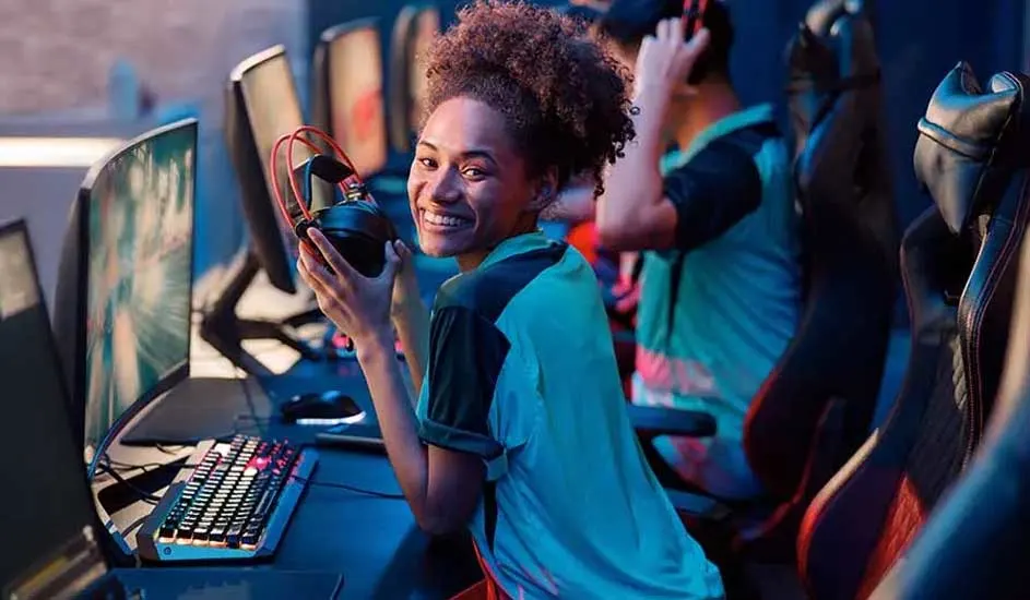 Une pièce avec des écrans d'ordinateur, des claviers et des chaises de jeu noires et rouges. Un casque noir et rouge est tenu à la main.