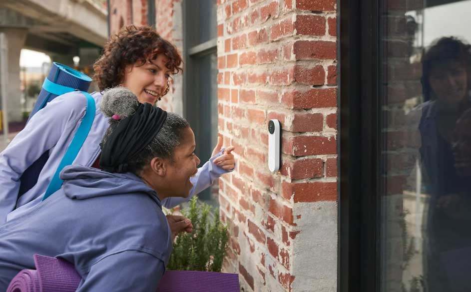 Twee vrouwen buiten wijzen naar een witte slimme deurbel aan een bakstenen muur, naast een donker raam.