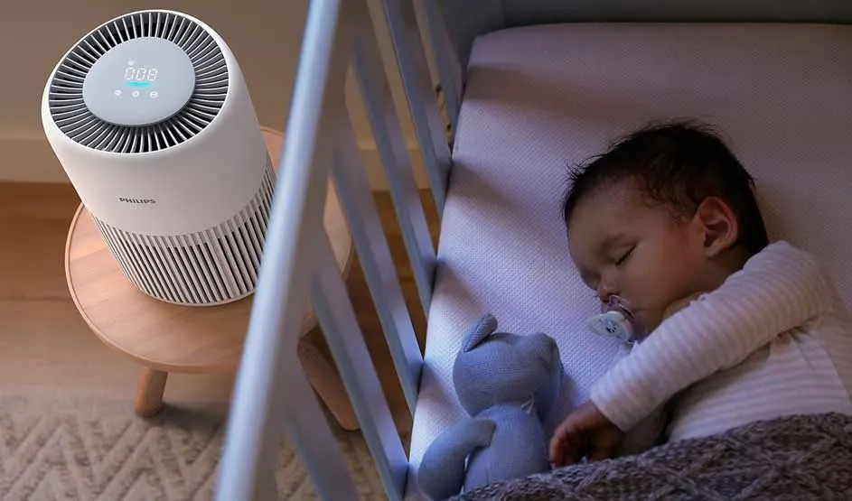 Baby slaapt rustig in een ledikant met knuffel, terwijl een Philips-luchtreiniger zachtjes draait op het krukje ernaast in de kinderkamer.