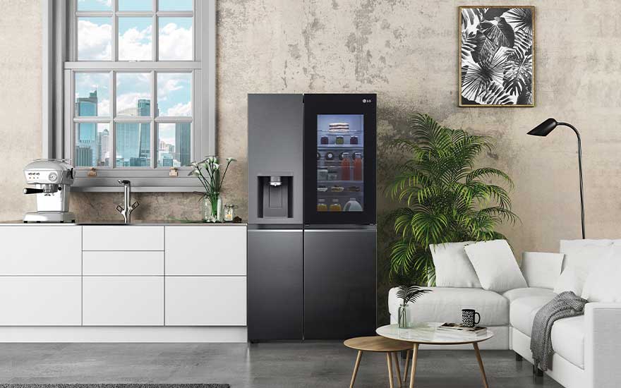 In een moderne keuken staat een grote zwarte LG koelkast met glazen deur, naast een wit aanrecht, planten en een comfortabele bank.