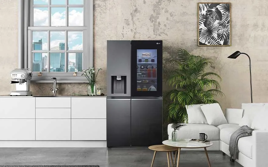 In een moderne keuken staat een grote zwarte LG koelkast met glazen deur, naast een wit aanrecht, planten en een comfortabele bank.